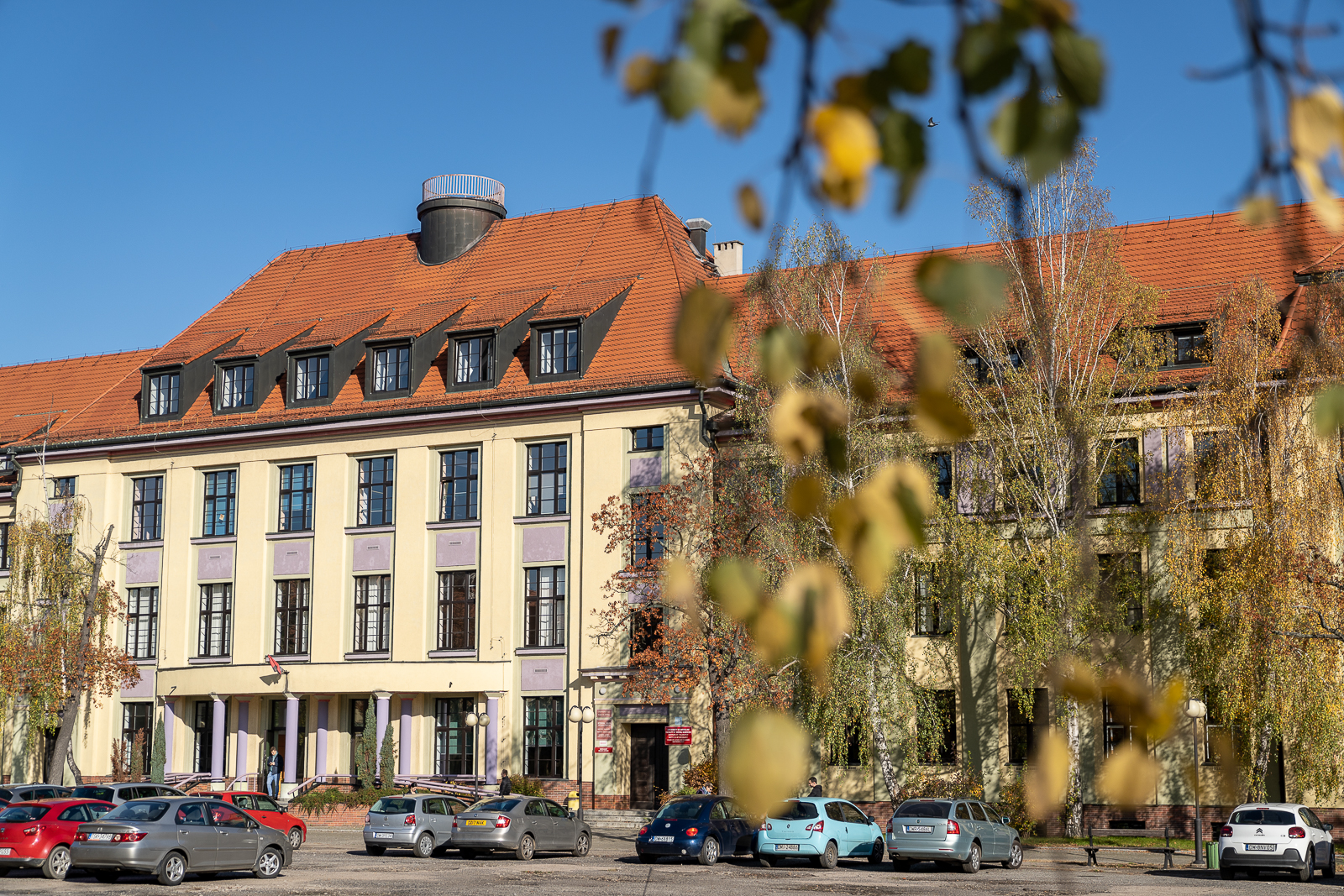 Żółty, duży budynek uniwersytecki, obok parking, na którym zaparkowane stoją samochody  