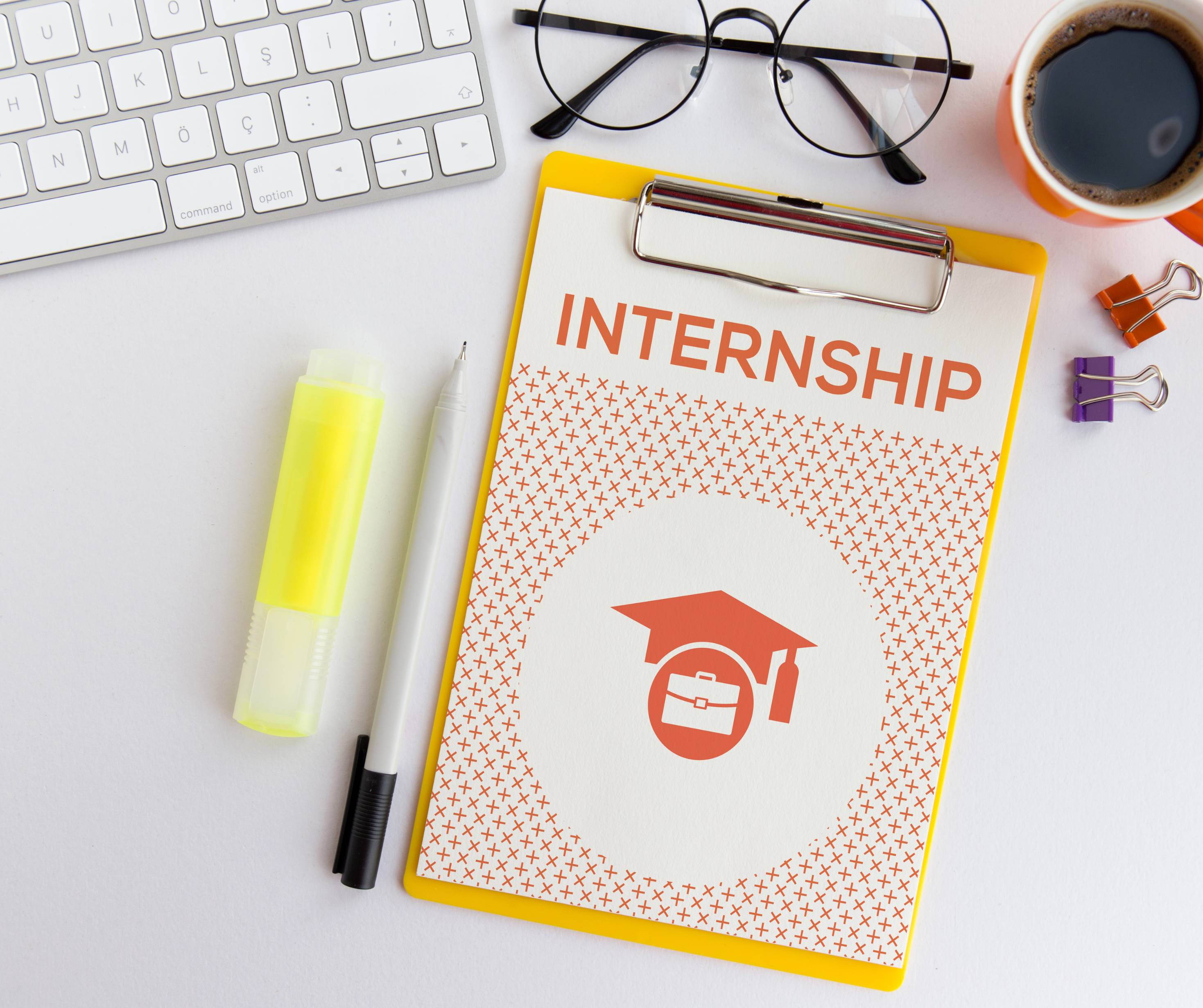 Kartka z napisem Internship leżąca na biurku. Obok stoi kubek kawy, okulary i długopisy