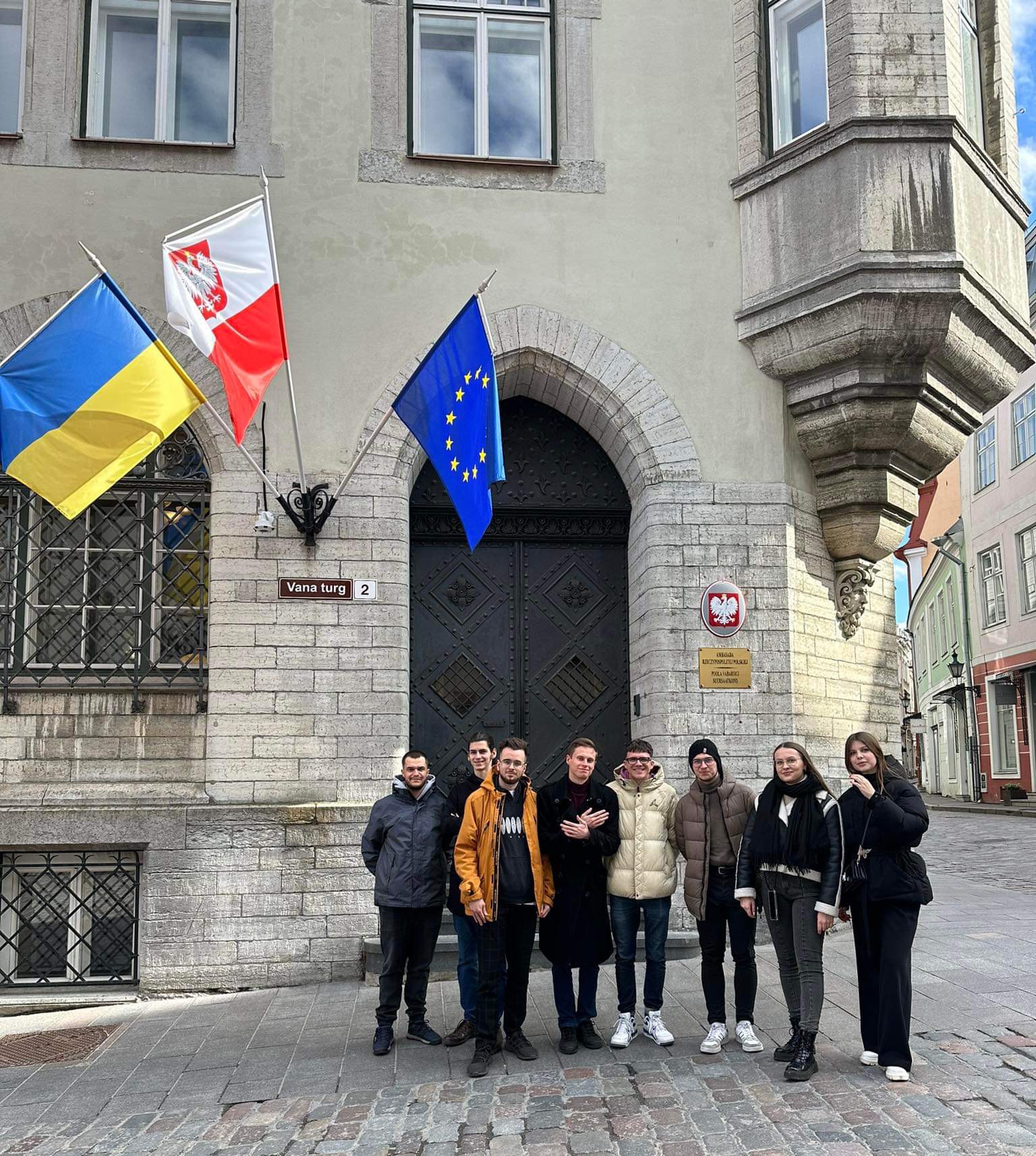 Eight people standing in front of a building with three flags in the background